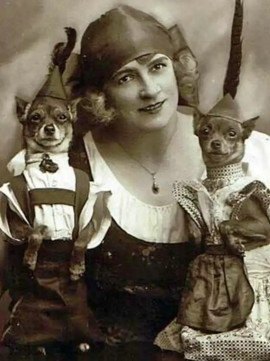 Vintage sepia portrait of a woman wearing a cloche-style hat, posed with two small dogs dressed in matching outfits, each perched at her shoulders, creating a whimsical anthropomorphic scene.