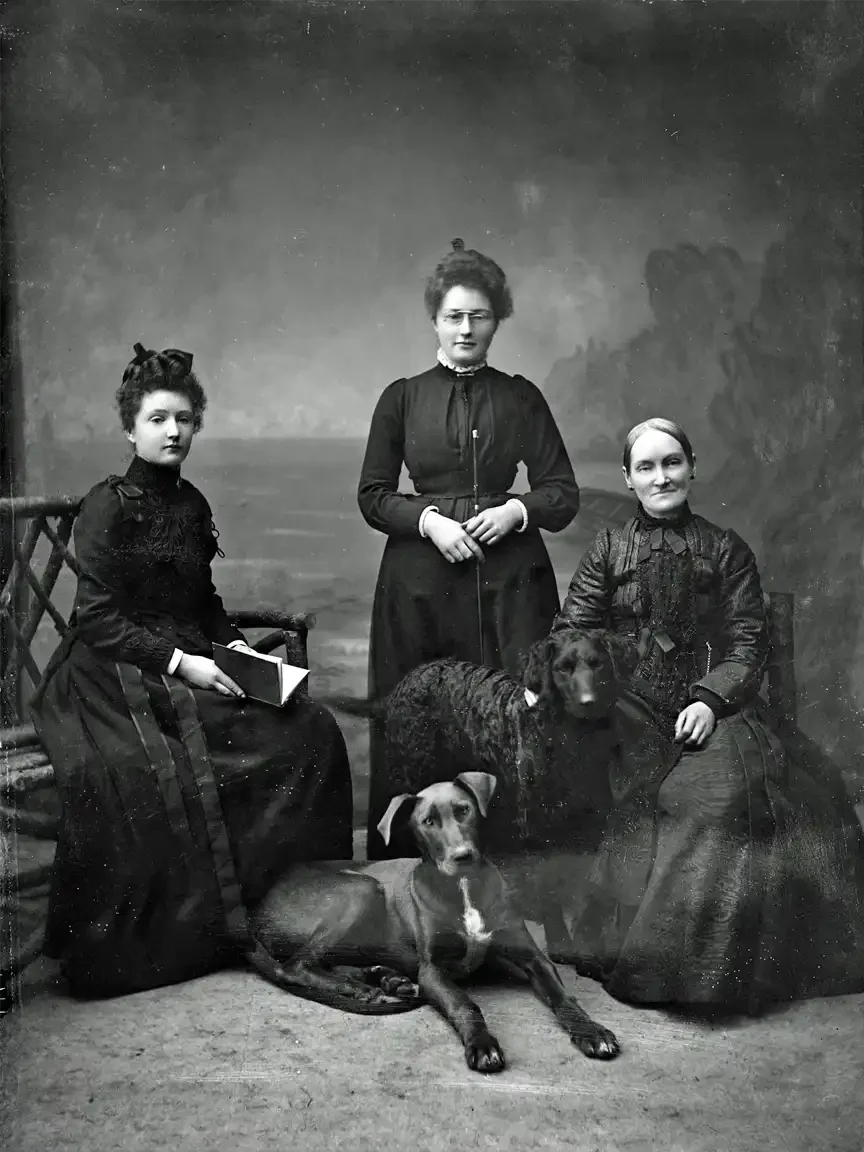 Three Victorian women pose in a studio portrait with two dogs—one seated, one lying at their feet—arranged formally against a painted backdrop in late 19th-century attire.