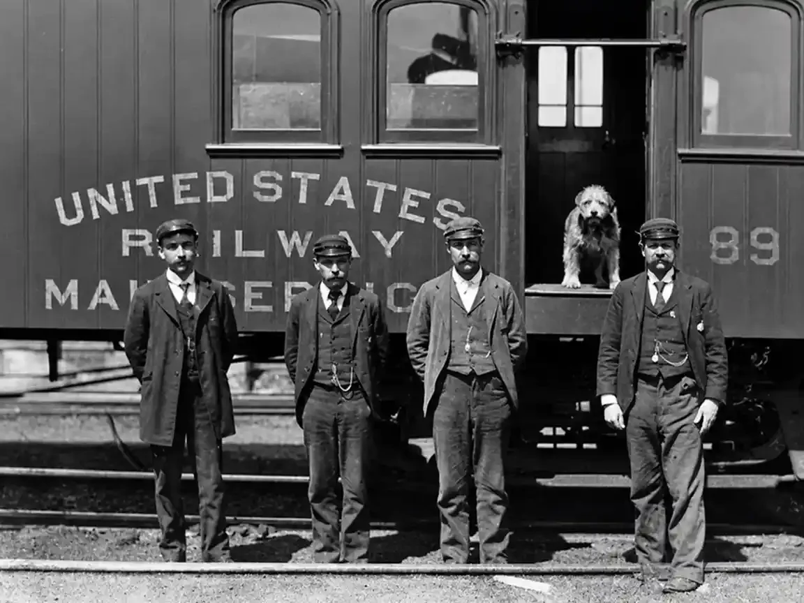 Four mail clerks in caps and waistcoats stand on the track before a Railway Post Office car; a small shaggy dog stands alone in the open doorway above them.