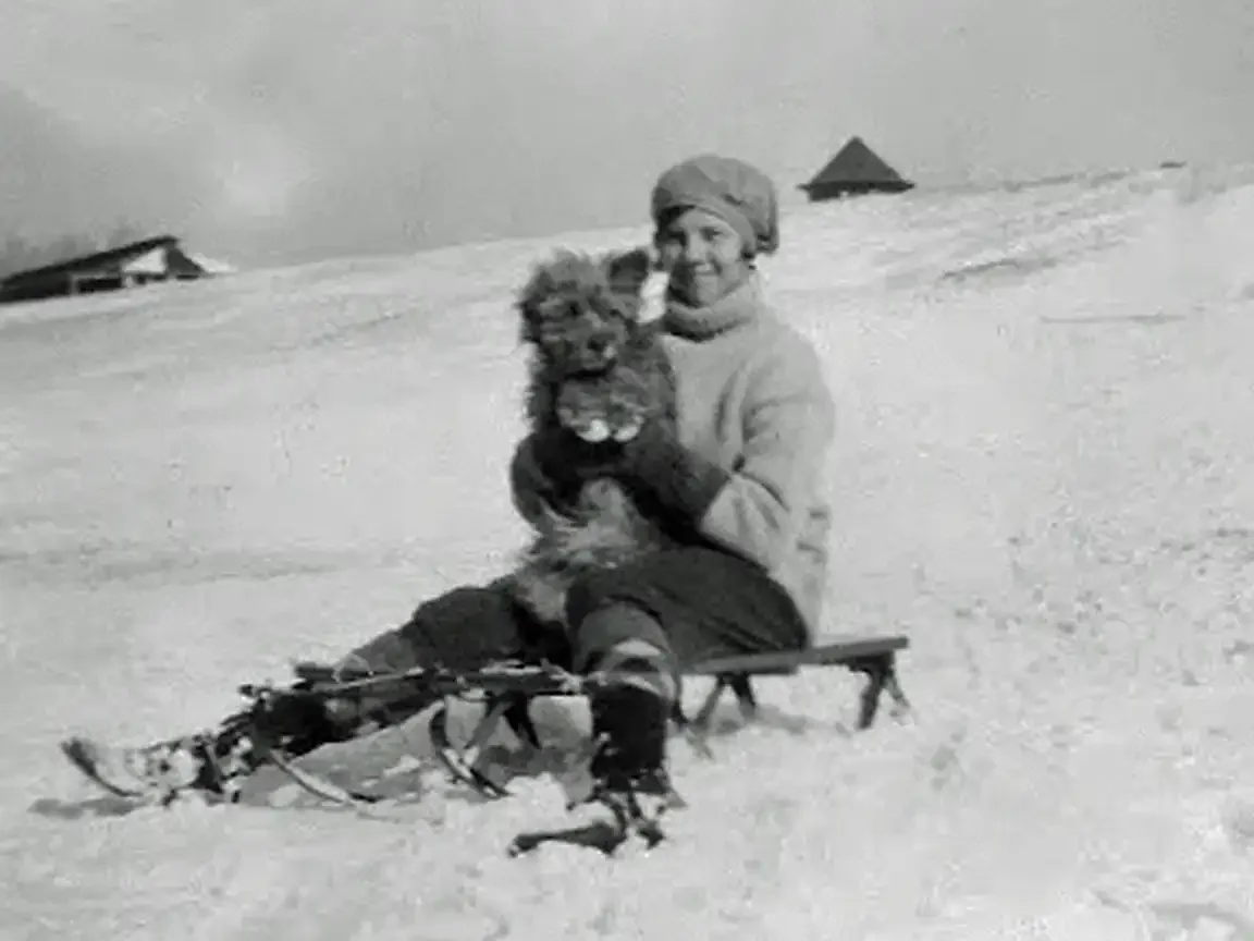 A smiling young woman holds a small shaggy terrier on a wooden sled in a snowy landscape, with military buildings visible in the background.