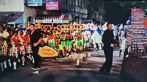 A festive street parade with colorful costumes and marching performers; two men in black are either side of a dog peeing in the middle of the street. Scene from James Bond movie Thunderball 1965