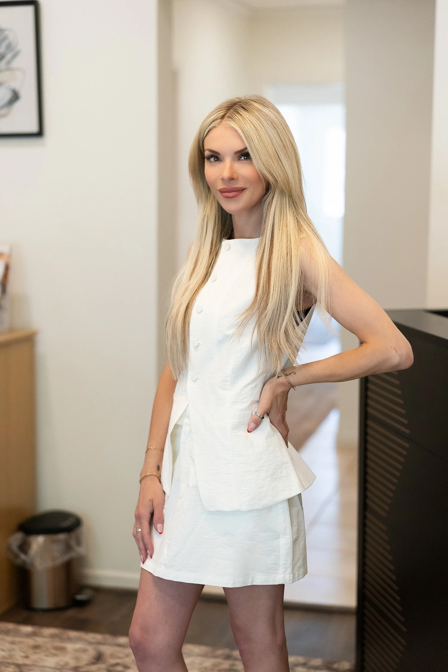 A woman with long blonde hair, wearing a white dress, standing indoors in a well-lit room, with her right hand on her hip, smiling slightly at the camera.
