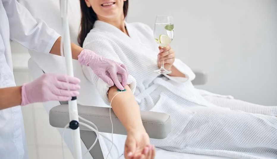 A woman in a hospital gown donating blood while sitting on a medical chair. A healthcare worker in pink gloves is drawing blood from her arm, and she is holding a glass of water with lemon slices.