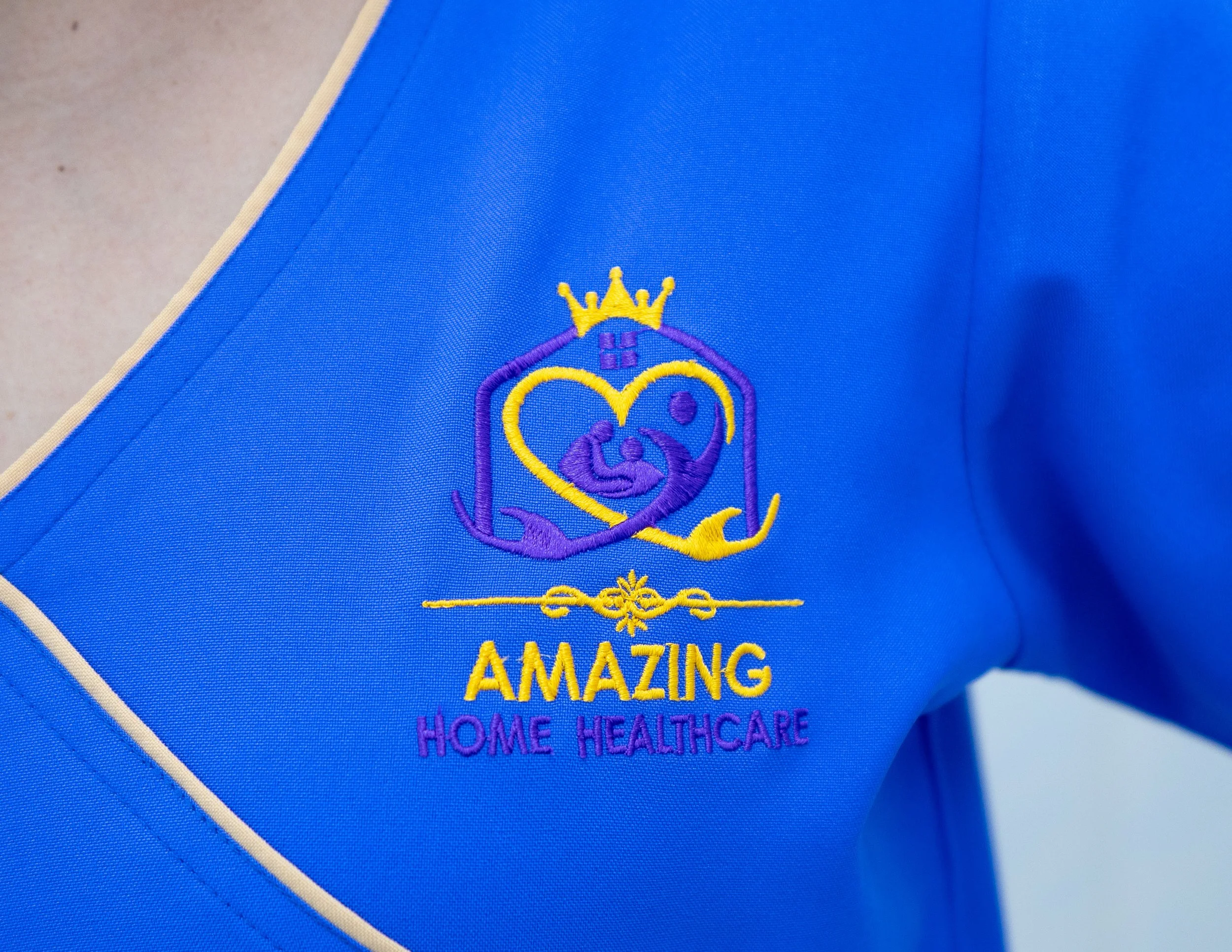 Blue uniform with embroidered logo and text reading 'AMAZING HOME HEALTHCARE', featuring a crown and heart design.