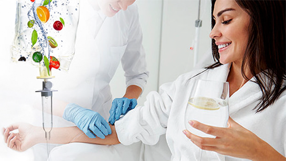 A woman in a white lab coat receiving an IV infusion of colorful fruit in a medical setting, while another person in a white coat and blue gloves tends to her arm and holds a glass of water.