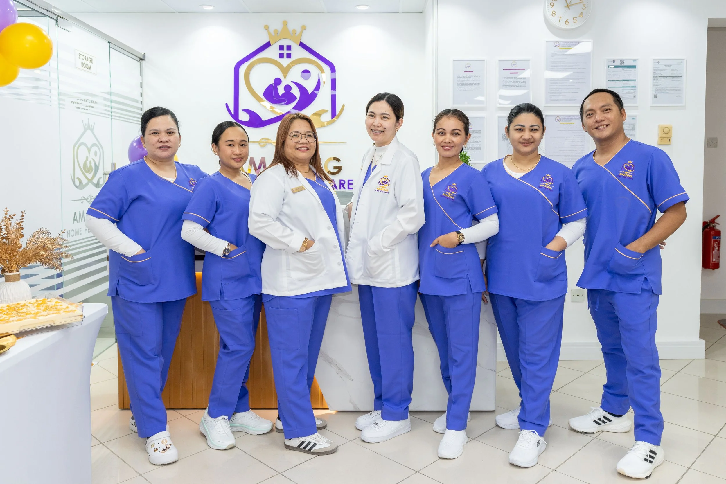 Group of seven healthcare professionals in blue scrubs and white coats standing together in a medical facility, smiling for the camera.