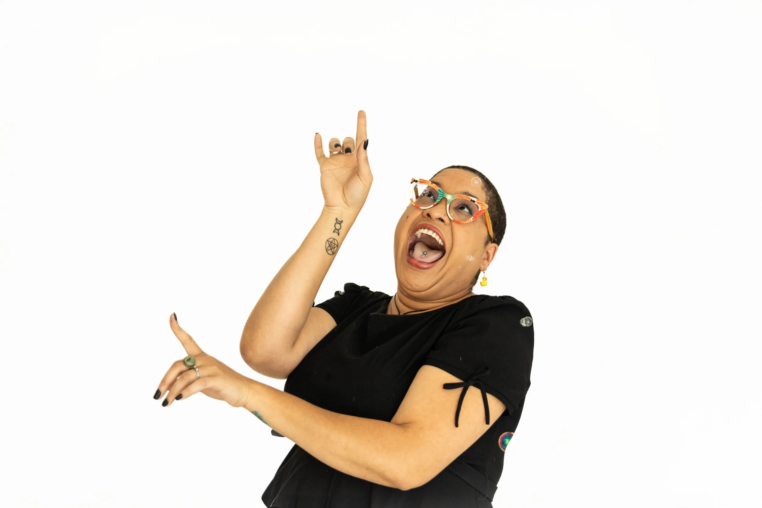 A joyful person with tattoos, wearing colorful glasses and black clothing, pointing upward with both hands and smiling widely.