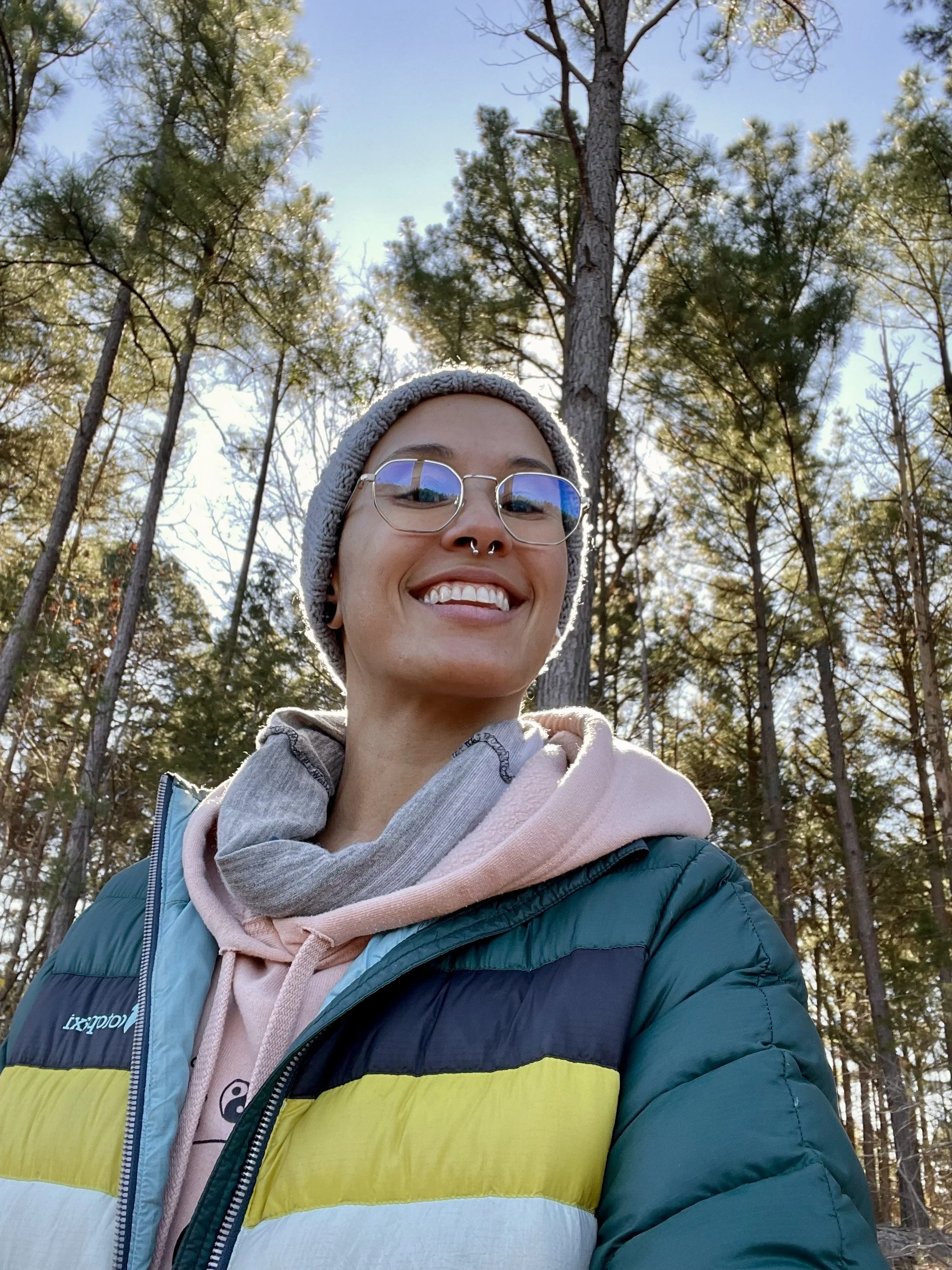 A person smiling outdoors in a forest, wearing glasses, a gray beanie, a hooded sweatshirt, and a puffy jacket.