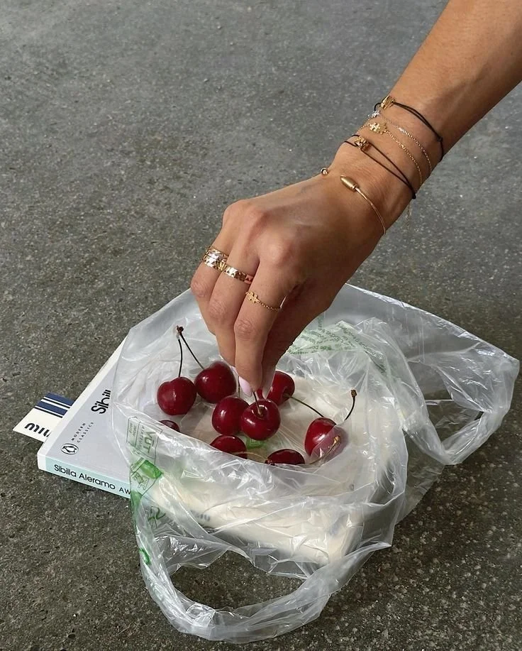 A person's hand reaching for cherries inside a plastic bag that contains a container of yogurt. The person is wearing multiple rings and bracelets.