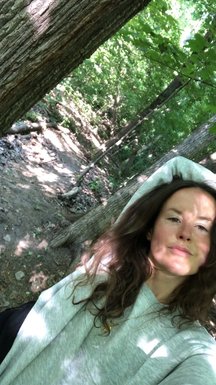 A woman with wavy brown hair taking a selfie in a wooded outdoor area with tall trees and green foliage.