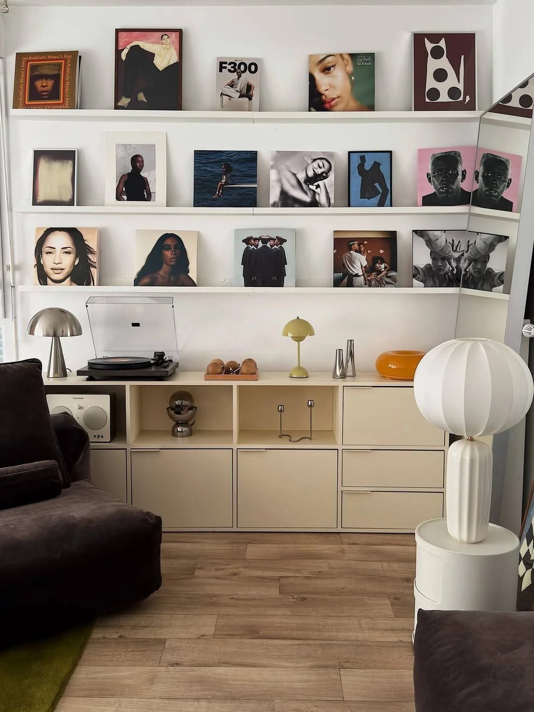 Wall-mounted shelves displaying various framed photographs and artwork, with a turntable on a white cabinet below, and a white lamp on a small round side table in a modern living room.