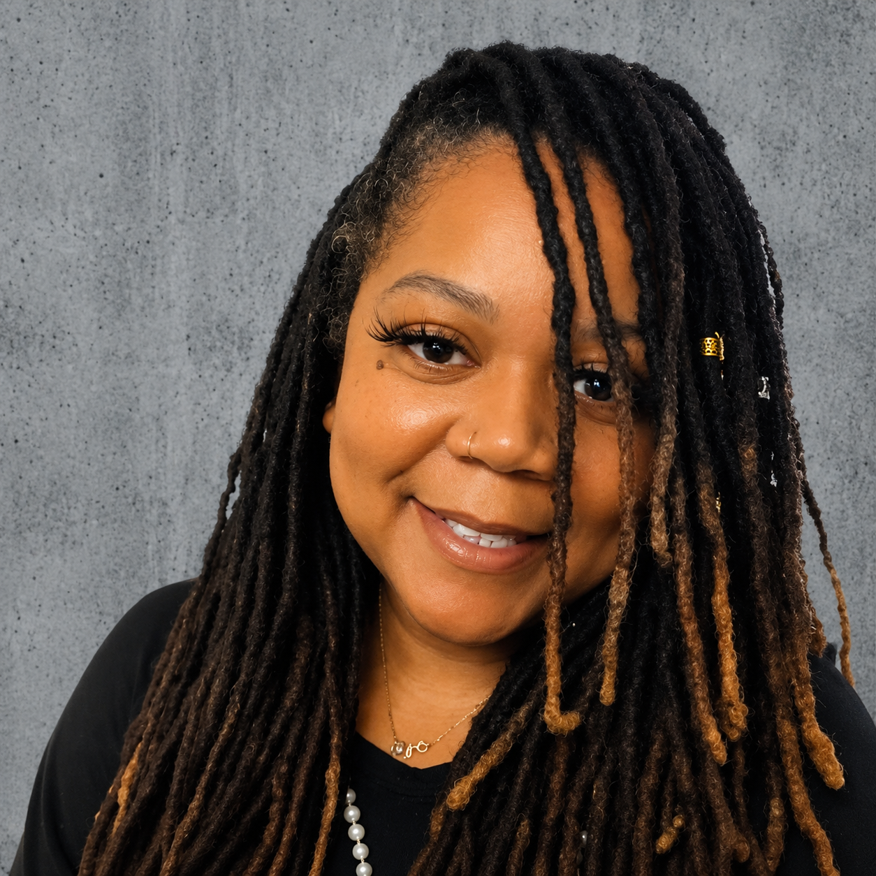 A woman with a warm smile, wearing gold and pearl jewelry, with long dark dreadlocks adorned with small decorative beads, standing in front of a textured gray wall.
