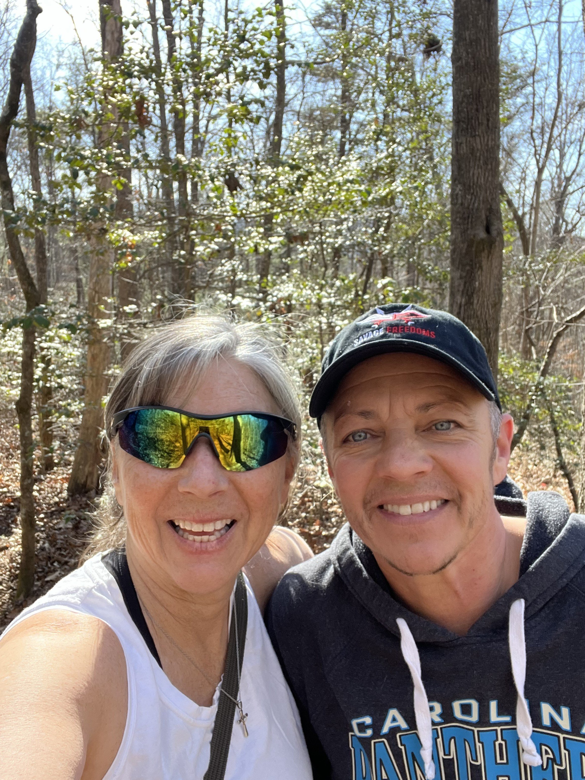 A selfie of a smiling woman with gray hair and sunglasses and a smiling man wearing a black cap and a Carolina Panthers hoodie, both outdoors in a wooded area on a sunny day.