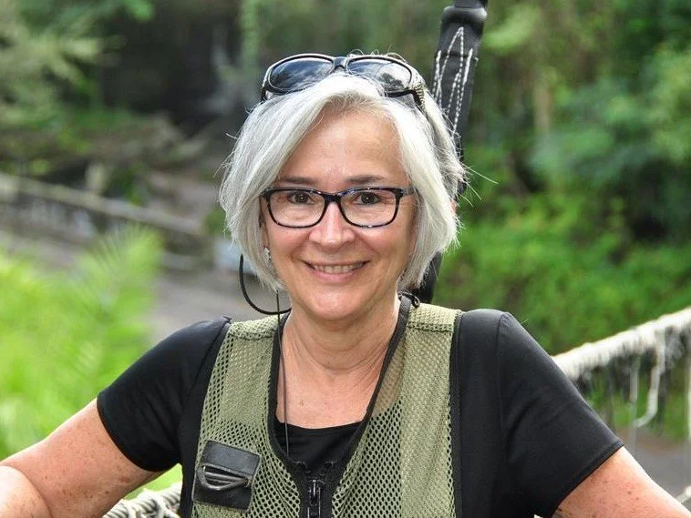 Smiling woman with short gray hair and glasses, outdoors in a forest setting, wearing a black t-shirt and a green hiking vest.
