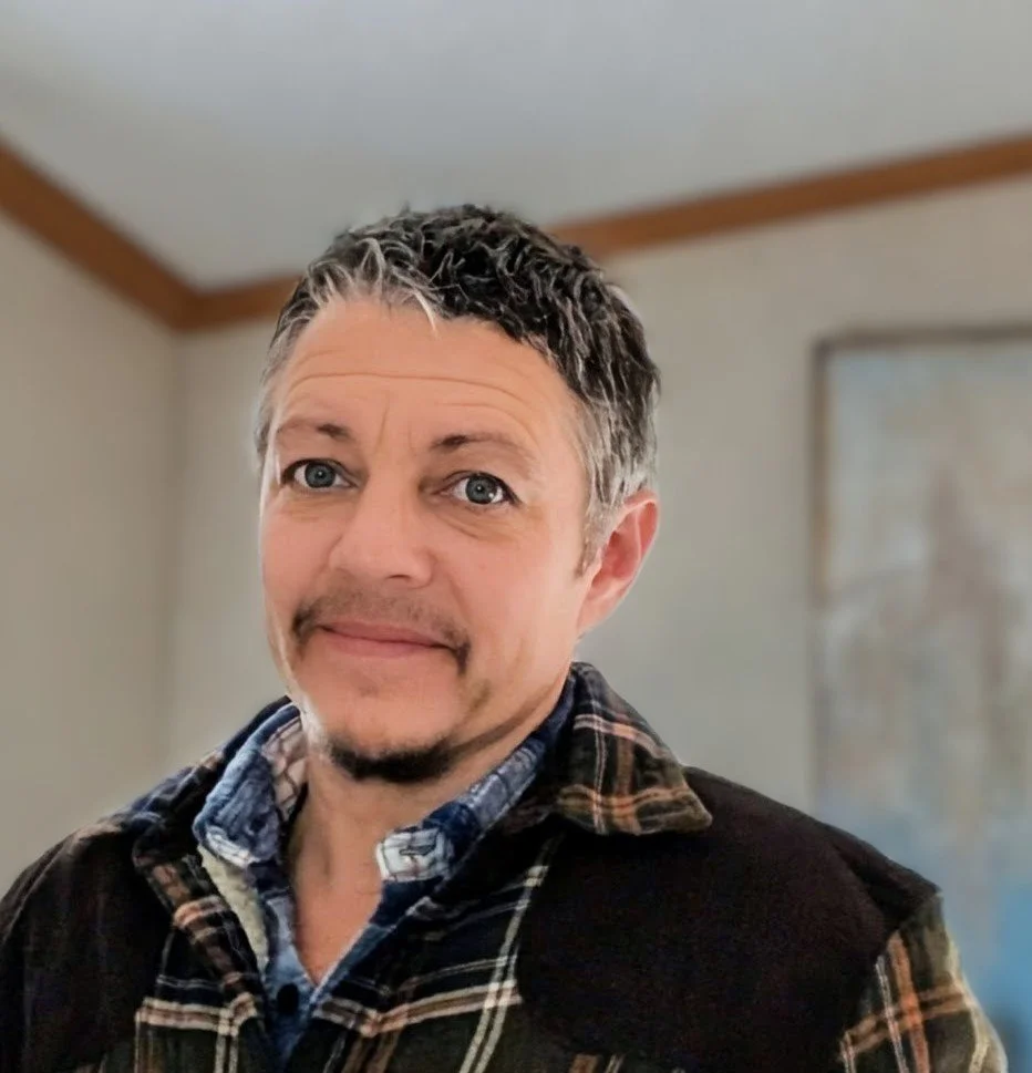 A middle-aged man with short, dark, and gray hair, smiling slightly, wearing a plaid flannel shirt and a dark jacket, standing indoors with light-colored walls and a framed map or picture in the background.