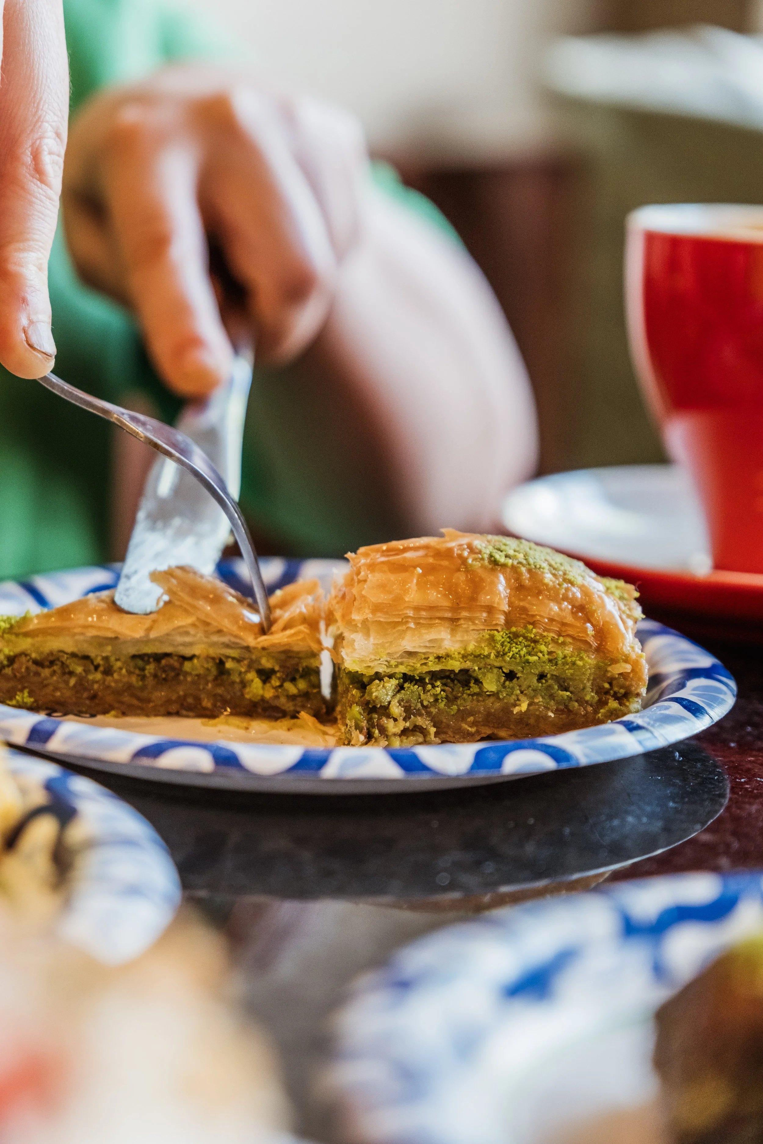 A person is serving a slice of pistachio and caramel pastry onto a blue and white paper plate, with a red mug and additional desserts in the background.