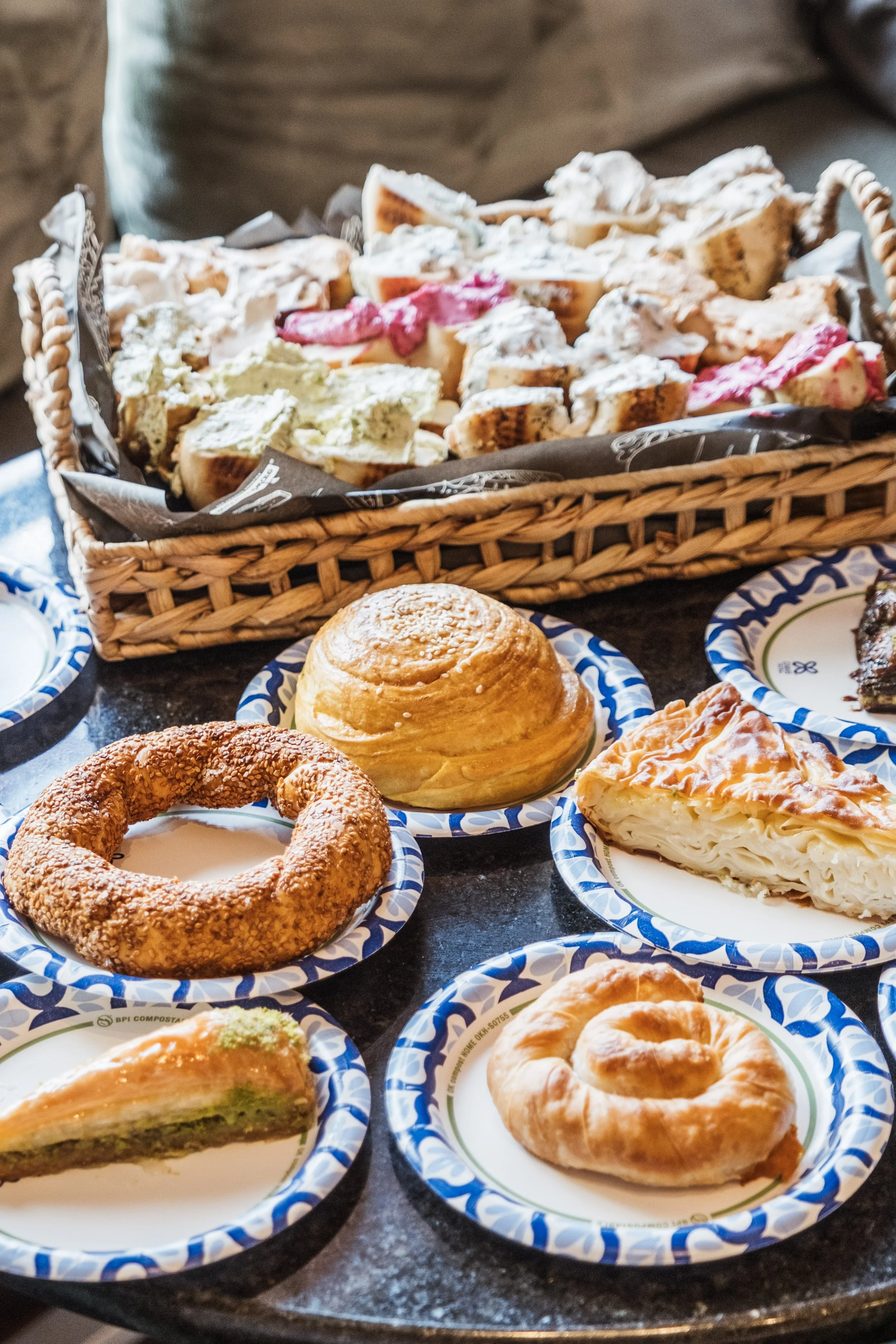 Assorted baked goods including croissants, a bagel with sesame seeds, a slice of green pistachio cake, and a cinnamon roll, with a basket of decorated cookies in the background.