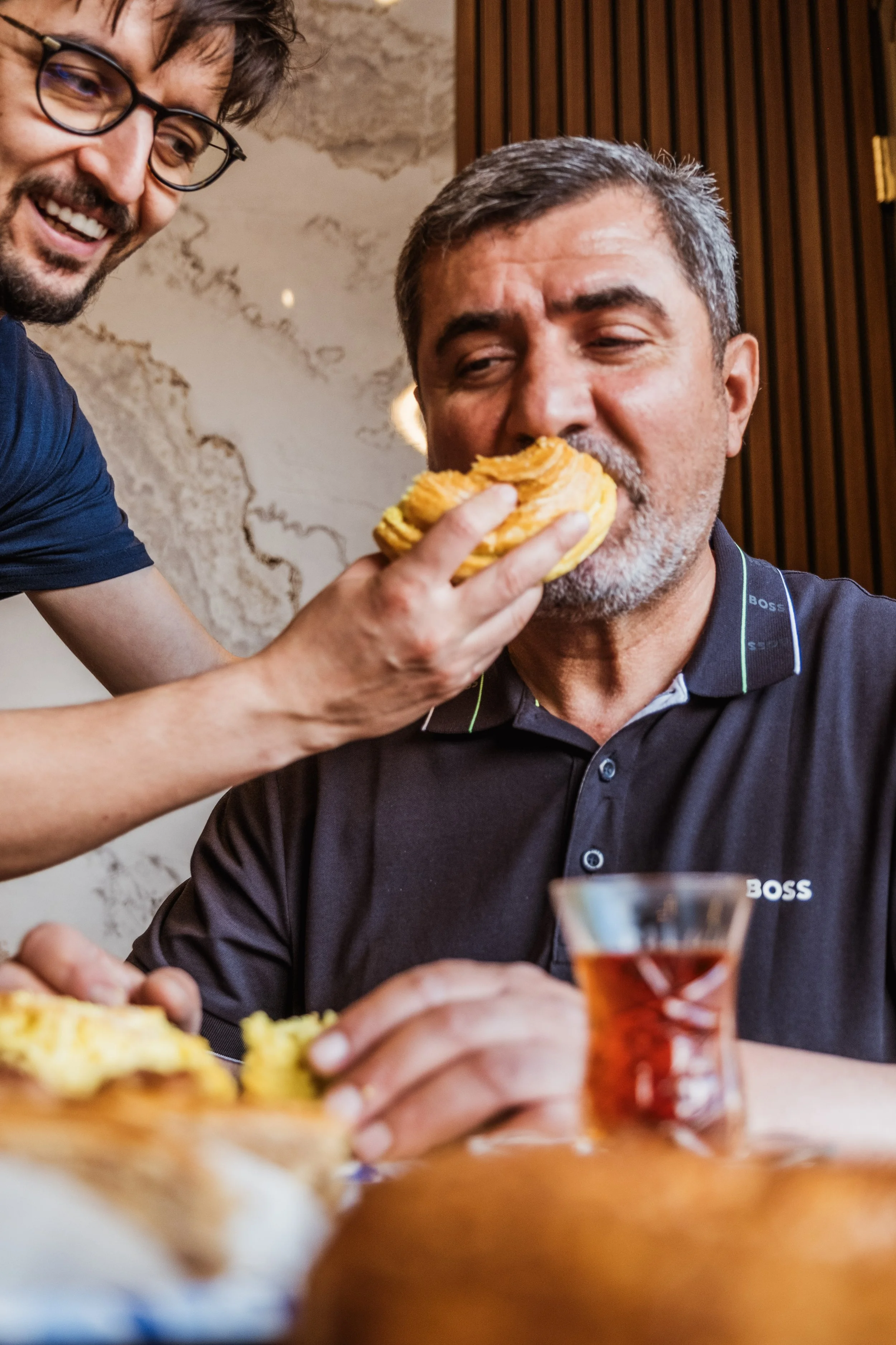 A man in a dark shirt with a smile on his face as another person feeds him a piece of pastry or bread in a restaurant setting.