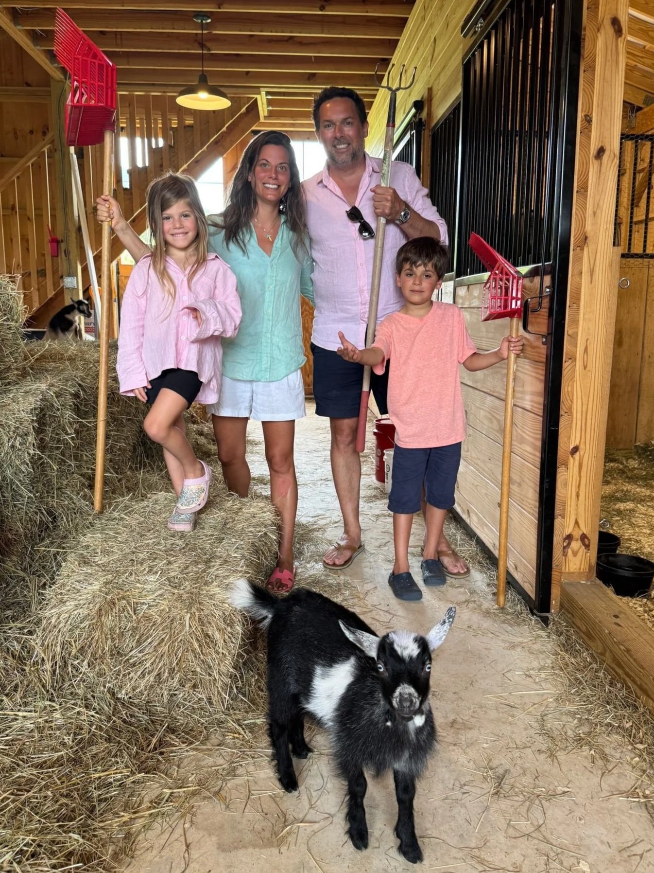 A family of four posing inside a barn, holding pitchforks. A young girl, woman, man, and young boy stand near hay bales, with a baby goat or kid in front of them. The barn has wooden walls and a black stall door.