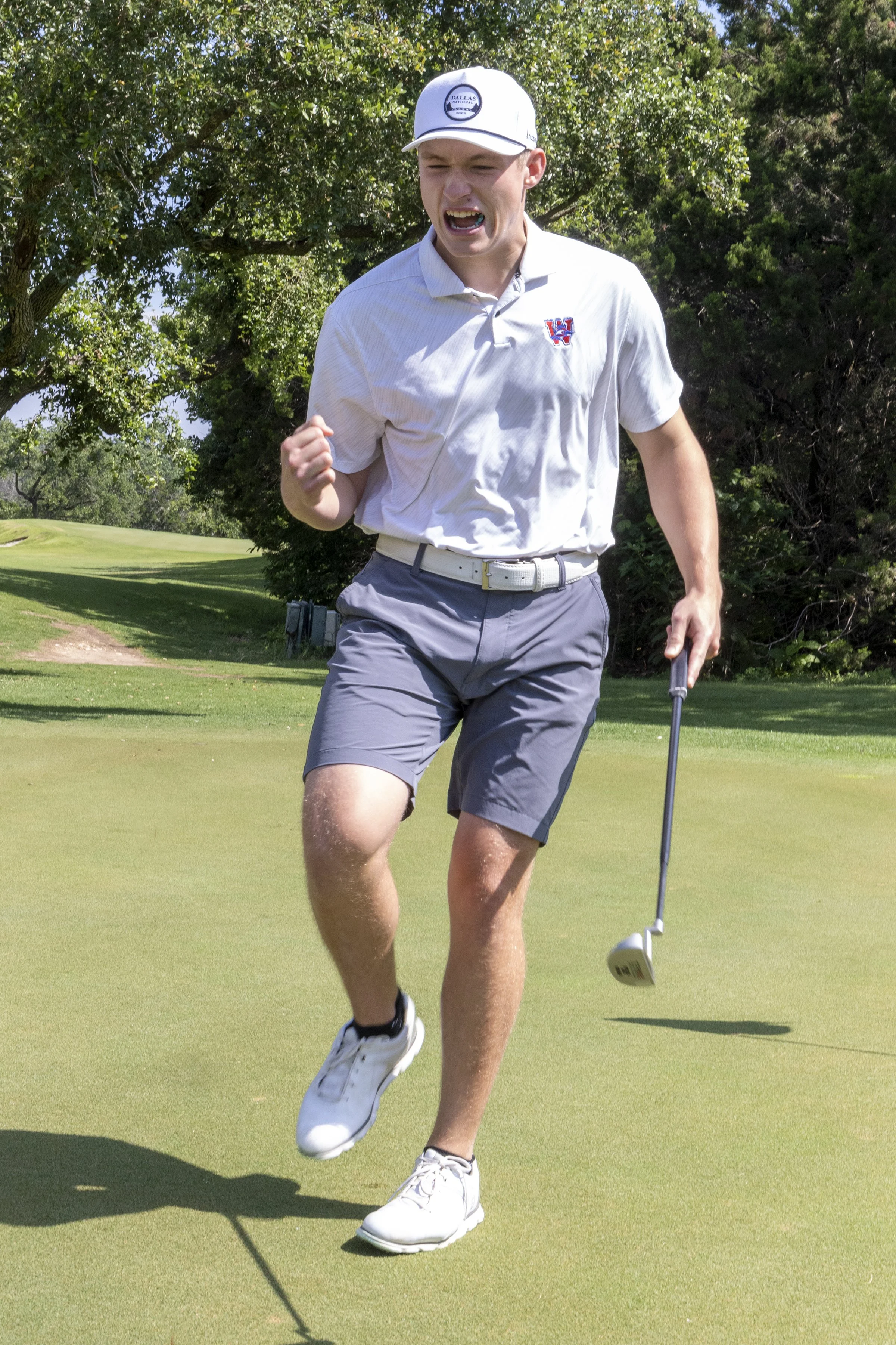 Golfer celebrating a successful golf shot on a green, wearing a white shirt, gray shorts, and a white cap, holding a putter in his right hand, with trees and a golf course in the background.