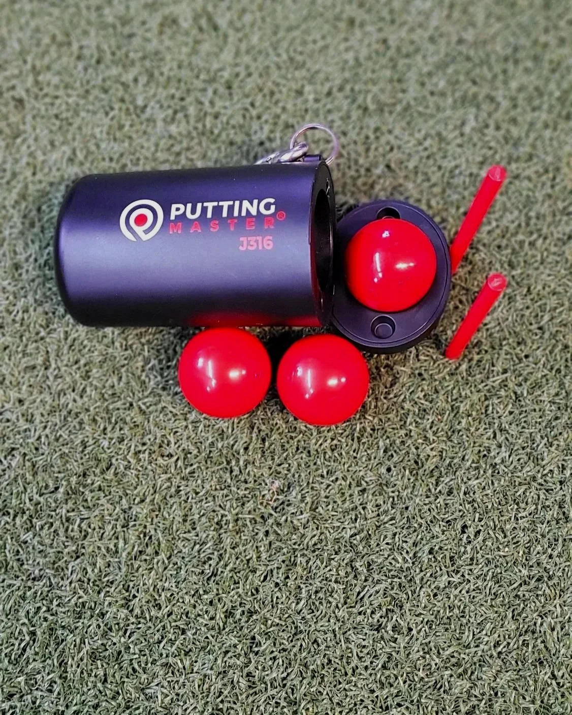 A golf putting training aid with a black container labeled 'Putting Master J316,' three red balls, and two red sticks, all placed on artificial green turf.