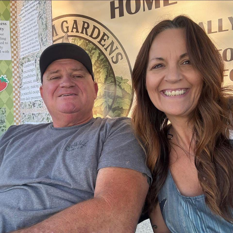 A man and woman are smiling in a photo, with the man wearing a black cap and the woman with long brown hair, in front of a backdrop with gardening-themed text and signs.