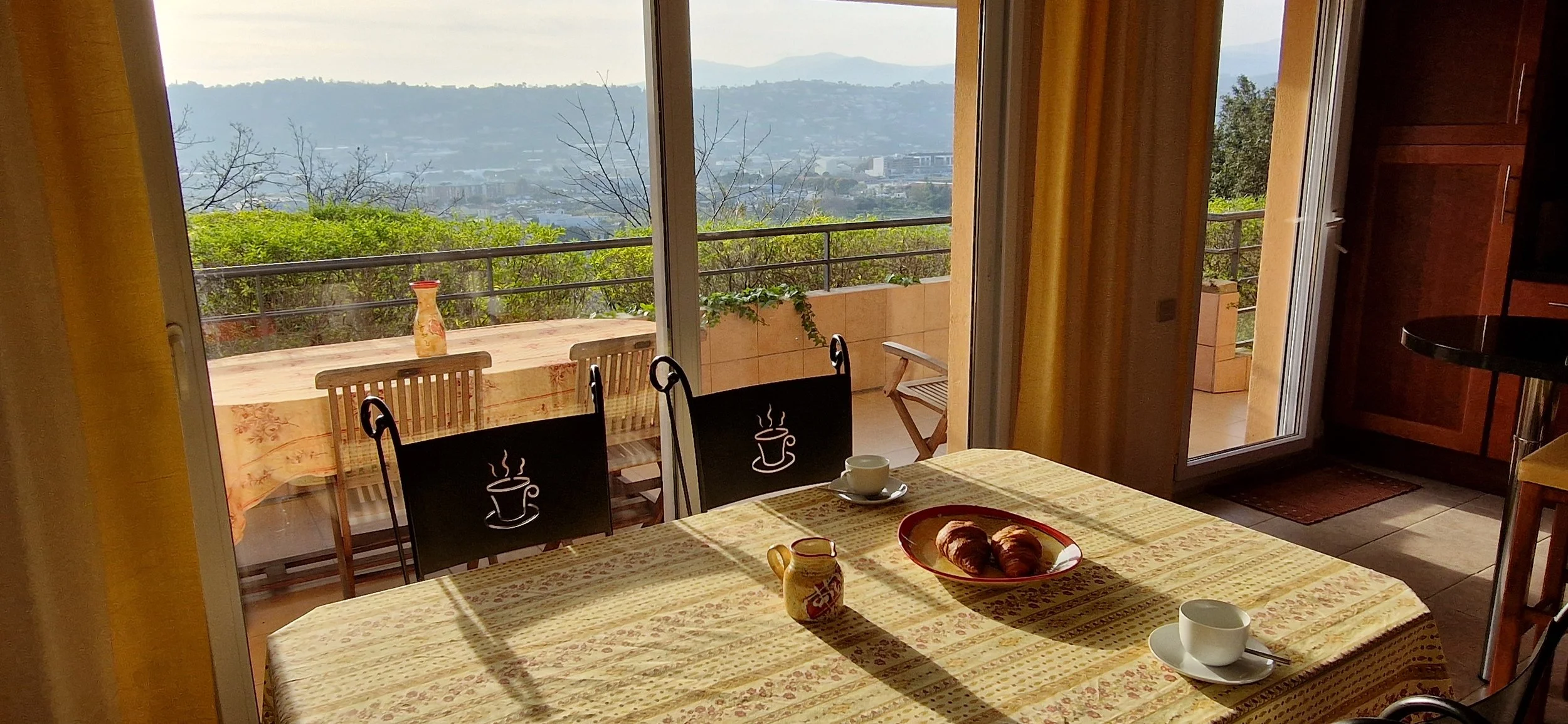 Aikataulu ja croissantit pöydällä, taustalla terassi ja kaupunkimaisema vuoristossa.