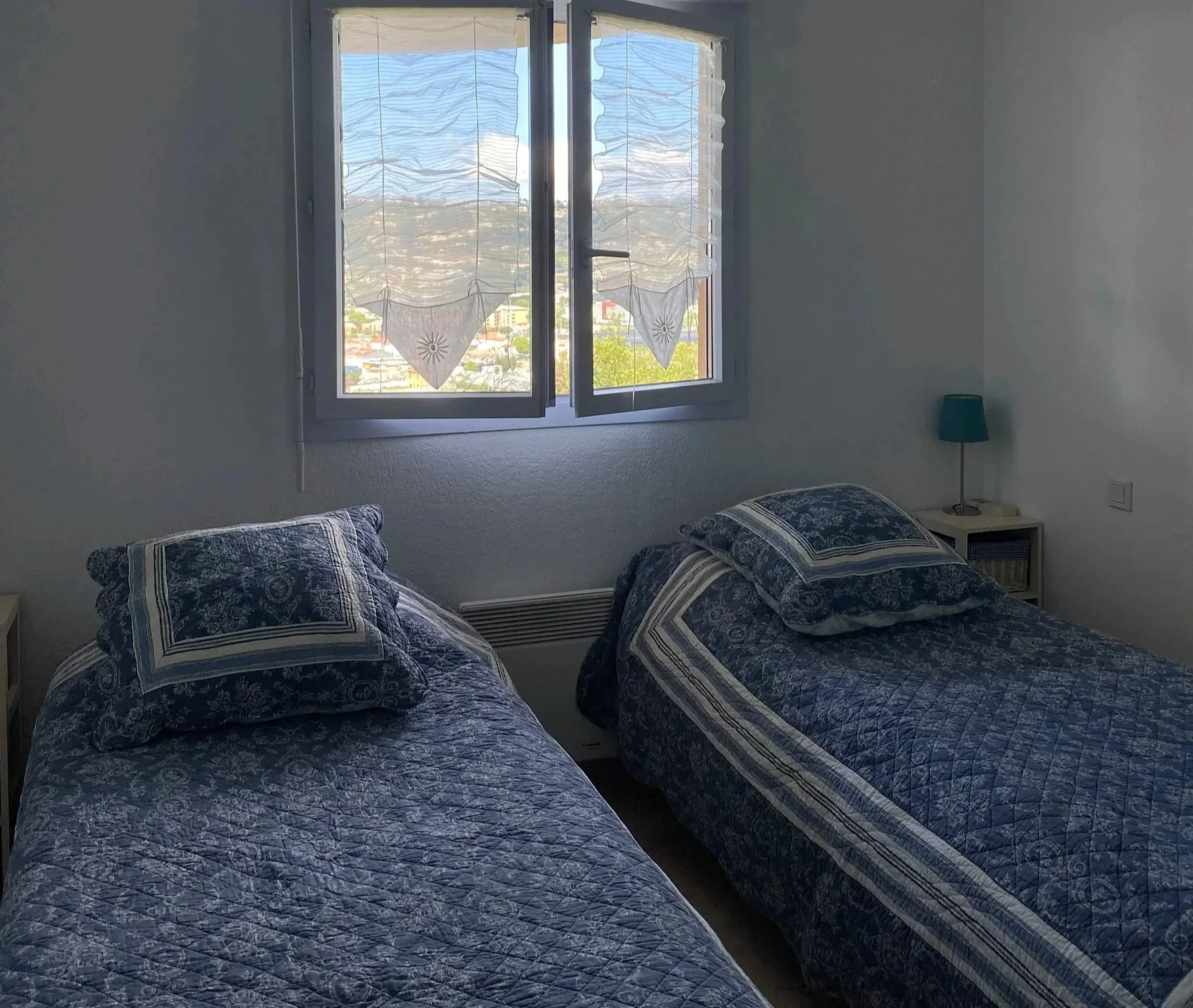 Kaks sänkyä, päivä- ja täysikokoinen, vuoratut sinisillä ja valkoisilla kankailla, huoneessa jossa on sivupöydät ja pieni sininen lamppu, ikkunassa valkoiset verhot, jotka näyttävät olevan osittain avoinna, ja näköala kaupunkiin vuorilla.