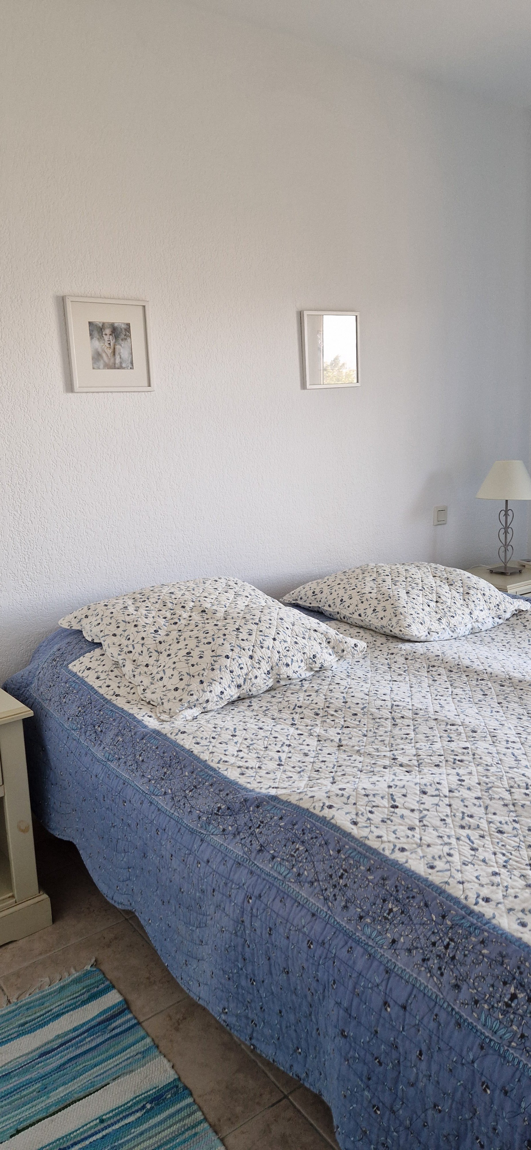 Sängyn päällä valkoinen päiväpeite ja kaksi koristetyynyä, seinällä kaksi taulua, yöpöydällä lamppu ja pieni kirja. Lattialla raitamatto, seinä on valkoinen ja vieressä ikkuna.