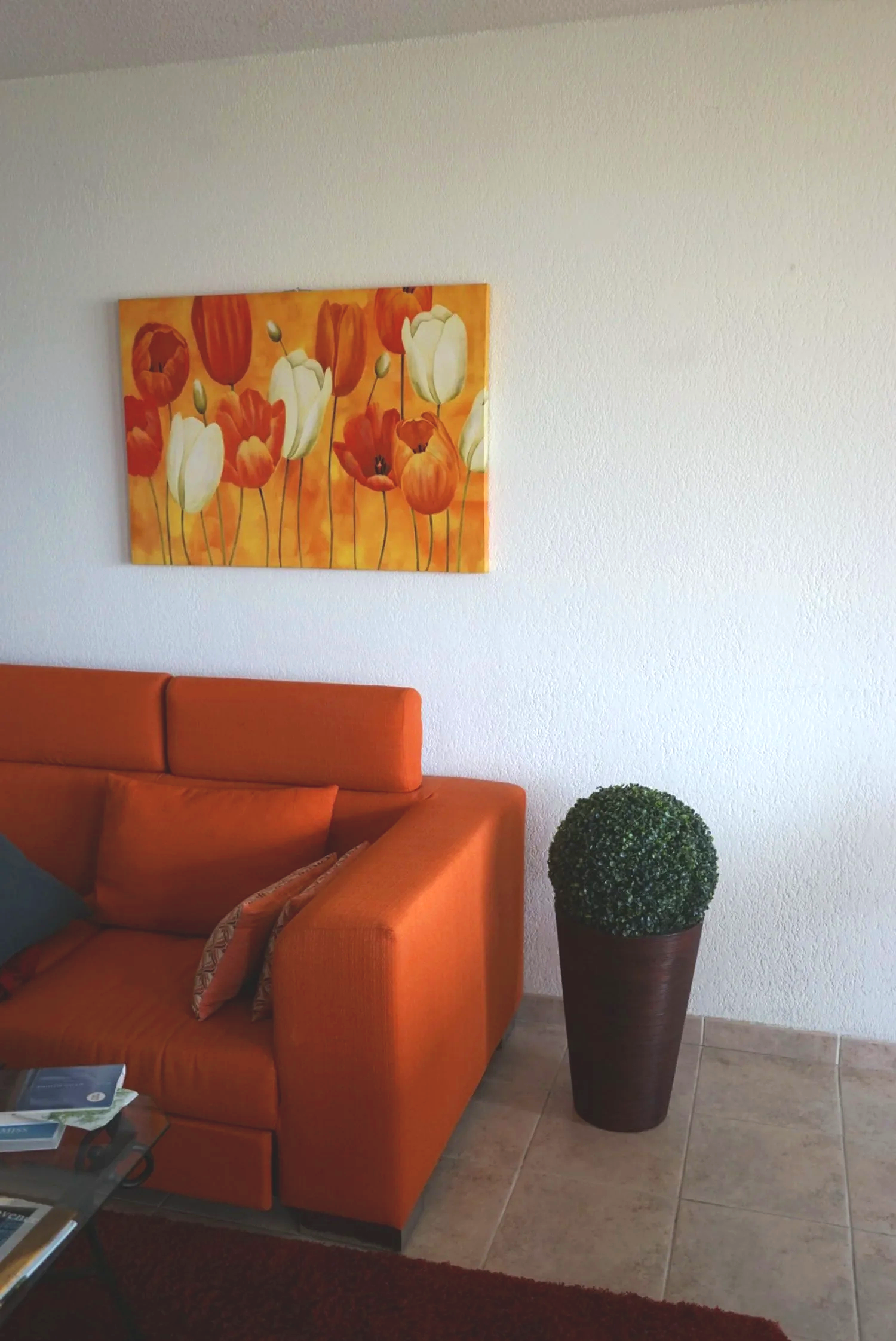 Oranssi sohva, taulu kukkakuviolla, pallo vihreää kasvia ruukussaan, osa olohuoneesta.