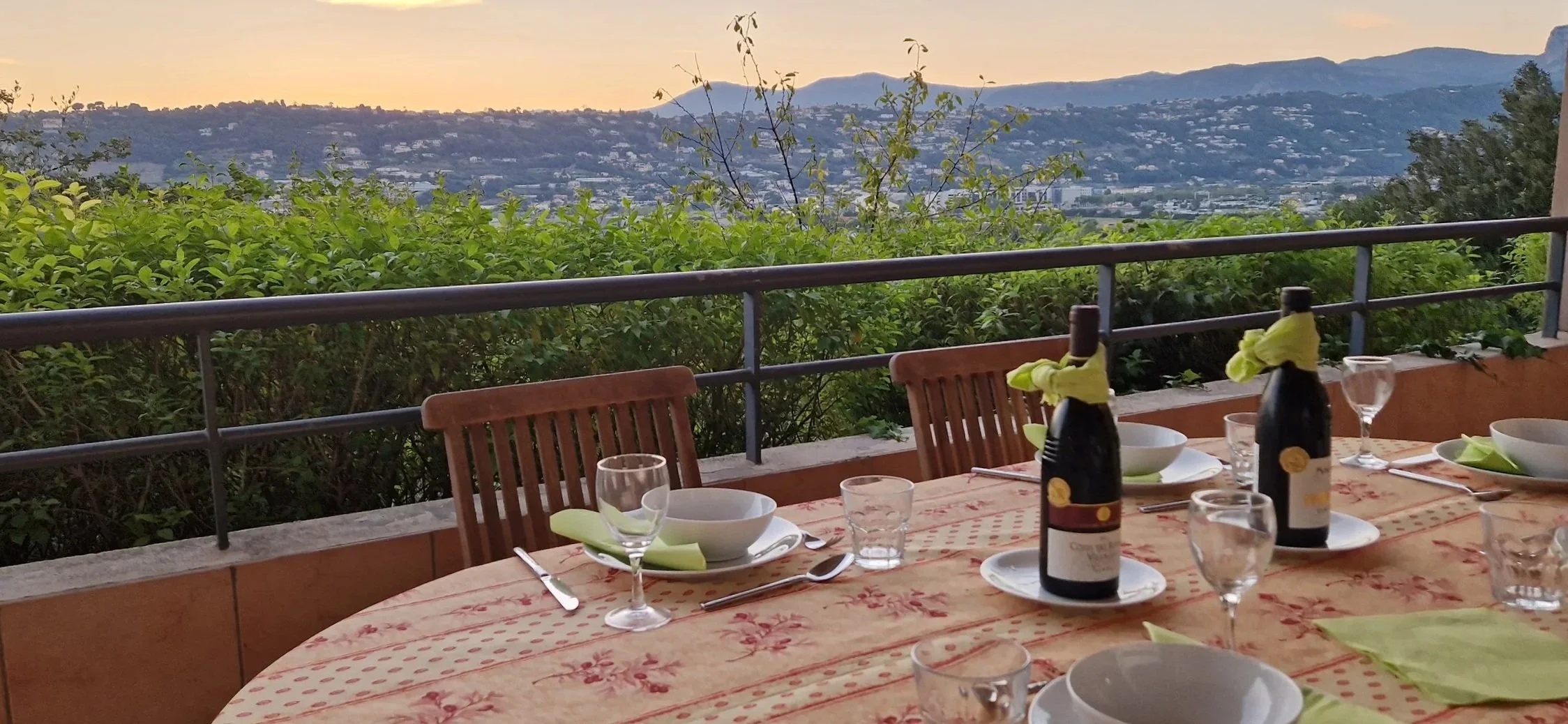 Ruokapöytä lasiovella, jonka takana vihreitä pensaita ja kaupunkinäkymä vuoristossa auringonlaskussa.