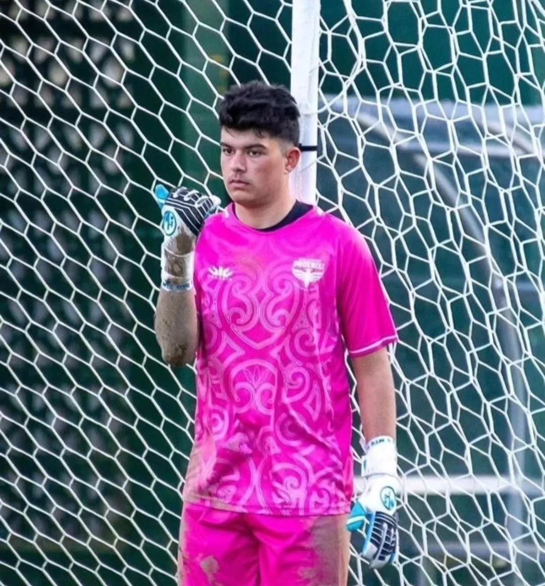 A young male soccer goalkeeper standing in front of a goal net, wearing a bright pink goalkeeper jersey and gloves, with a serious expression on his face.