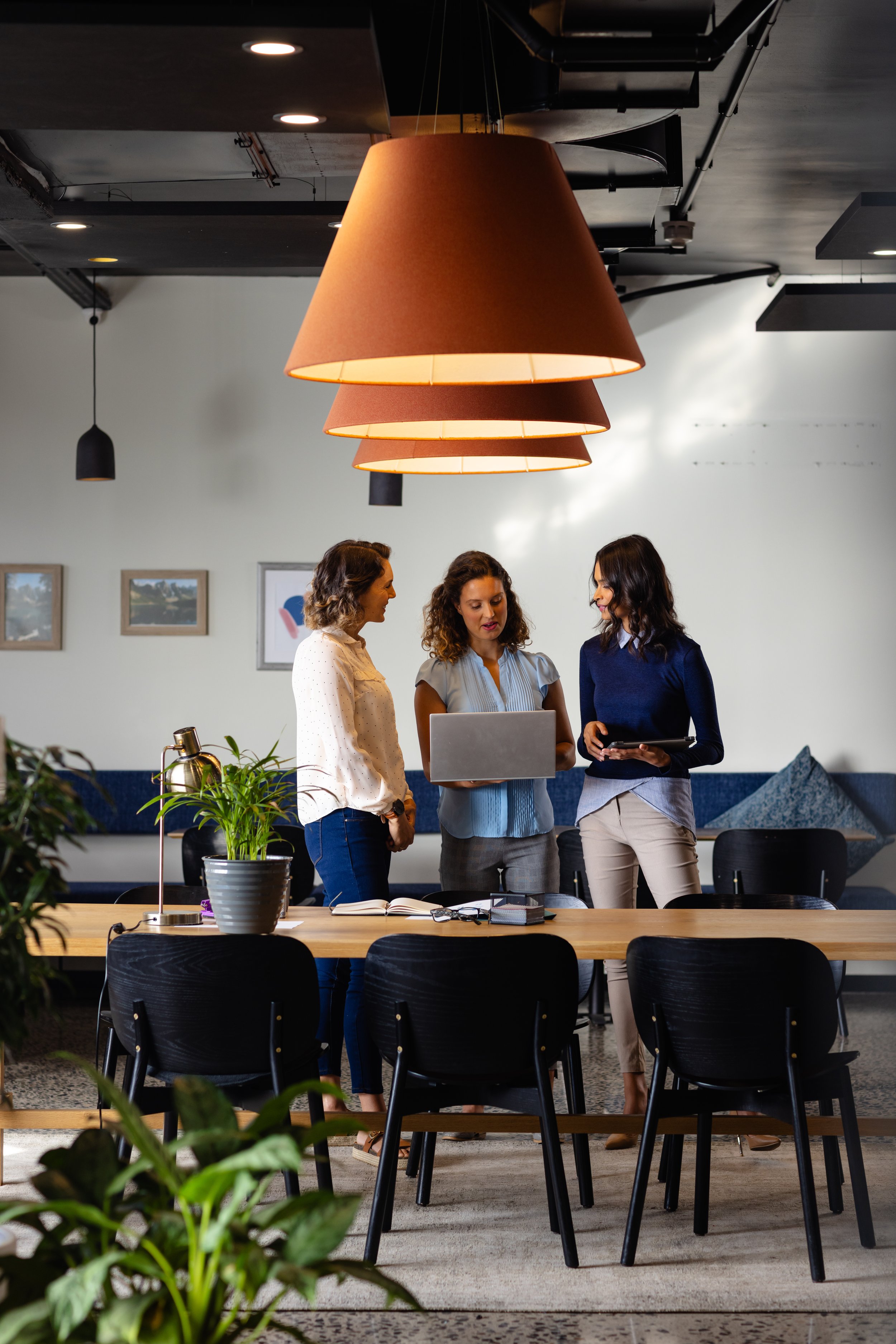 Three women standing around a table in a modern office, discussing work while looking at laptops and a tablet.