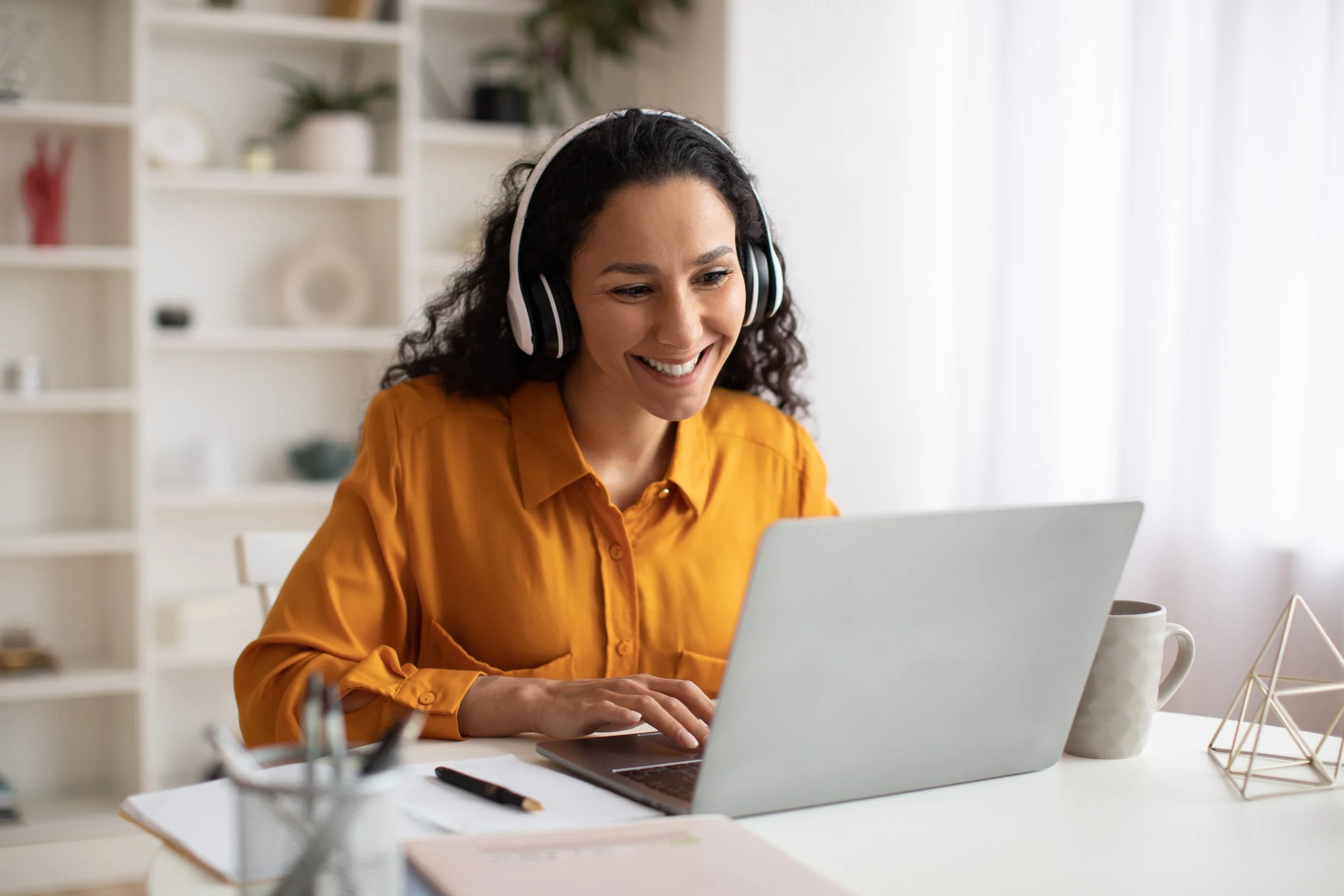 Smiling woman in a mustard yellow blouse wearing black and white headphones, working on a silver laptop at a white desk in a bright room.