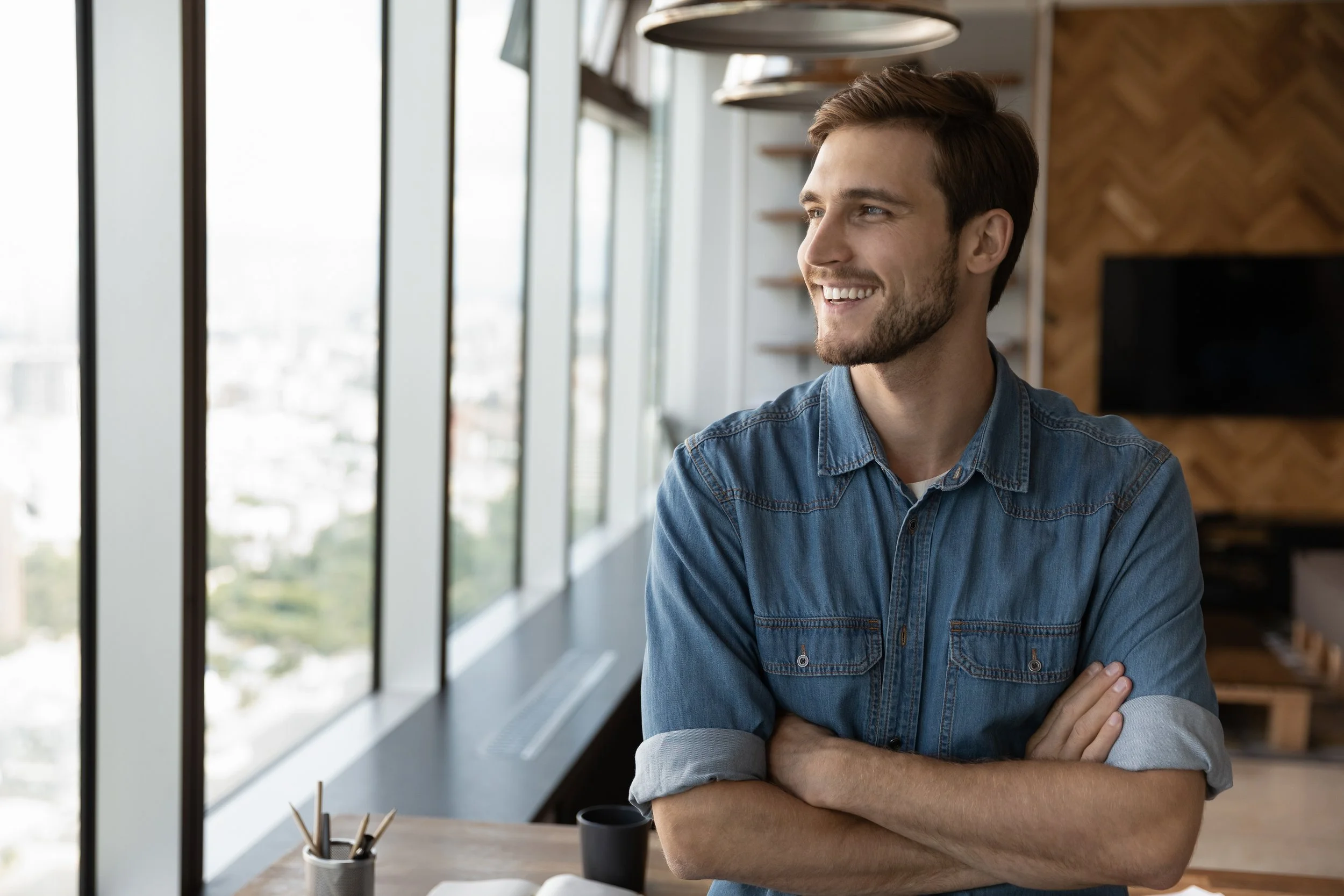 A young man with a beard and short brown hair, wearing a denim shirt, smiling while sitting with arms crossed in a modern, brightly lit office space with large windows.