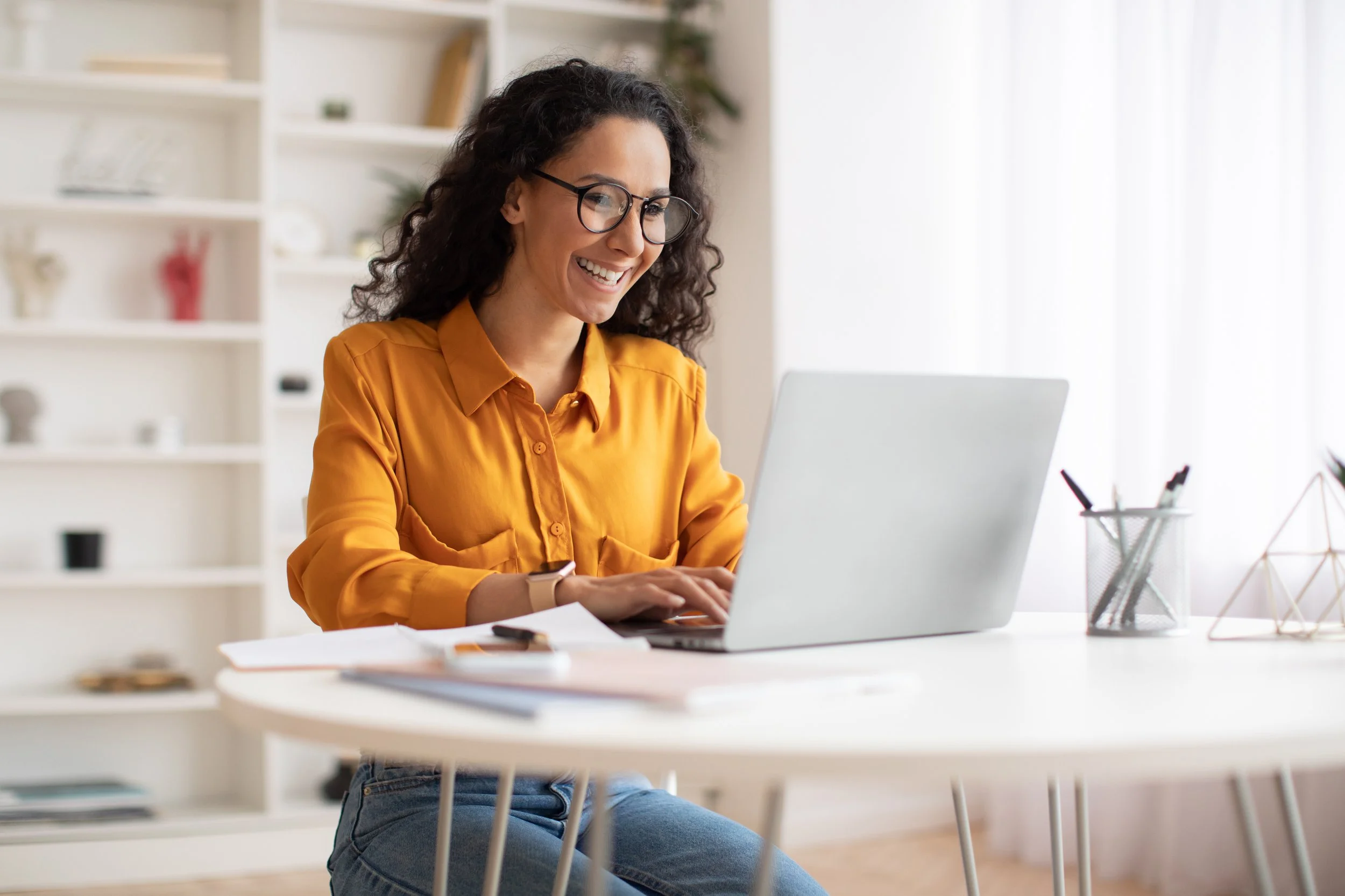 A woman with curly dark hair, glasses, and a mustard yellow shirt working on a laptop at a white table, smiling.