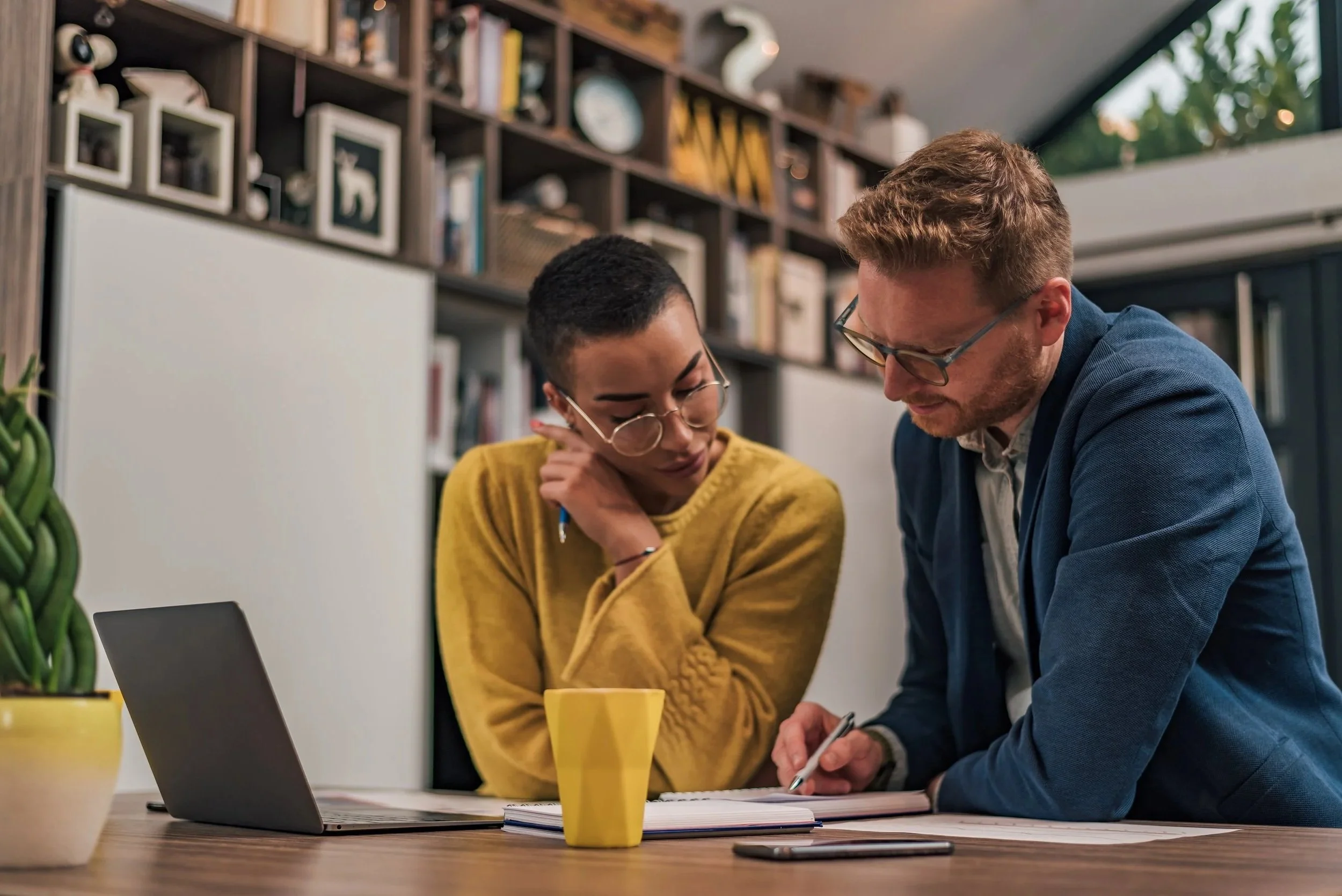 Two people, one woman and one man, working together at a desk in a modern office. The woman is wearing a yellow sweater and glasses, and the man is wearing glasses and a blue blazer. They are looking at documents on the table, with a laptop, a yellow mug, and a smartphone on the desk.
