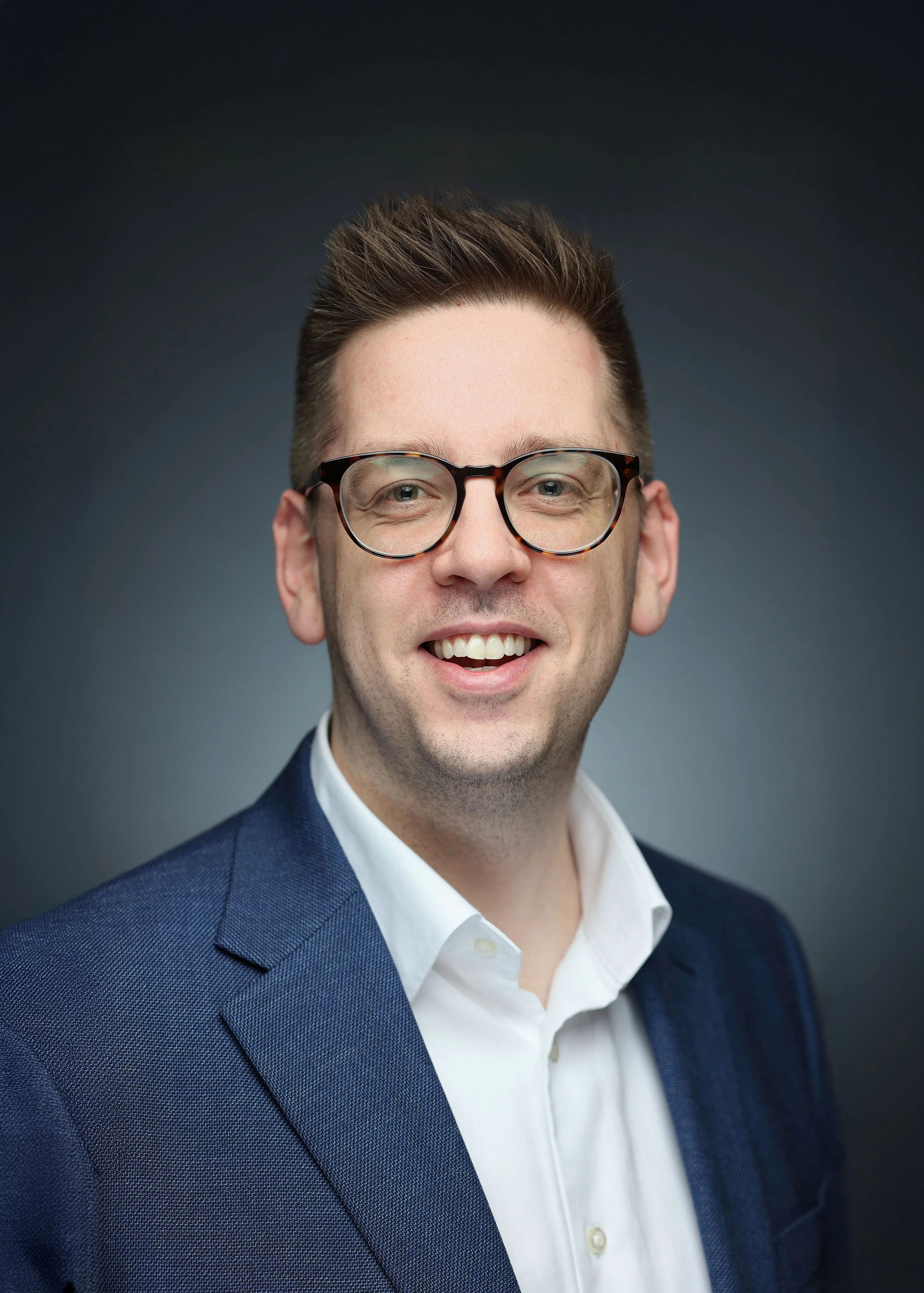 Professional portrait of a smiling man with short brown hair, wearing black-rimmed glasses, a white button-down shirt, and a dark blue blazer, against a gradient dark gray background.