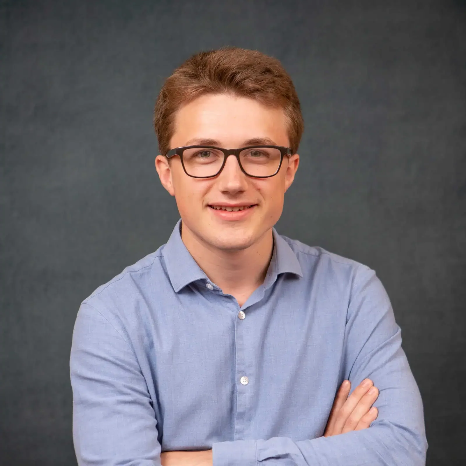 A young man with light brown hair, wearing glasses and a blue button-up shirt, smiling with arms crossed against a dark gray background.