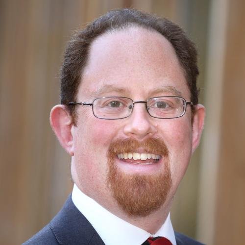 A man with glasses, a beard, and a mustache smiling, wearing a suit and tie.