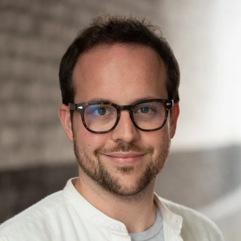 A young man with short brown hair, glasses, and a beard, smiling at the camera. He's wearing a white shirt and is indoors with a blurred background.