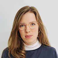 Portrait of a young woman with light brown hair, wearing glasses, a white turtleneck, and a blue blazer, against a plain gray background.