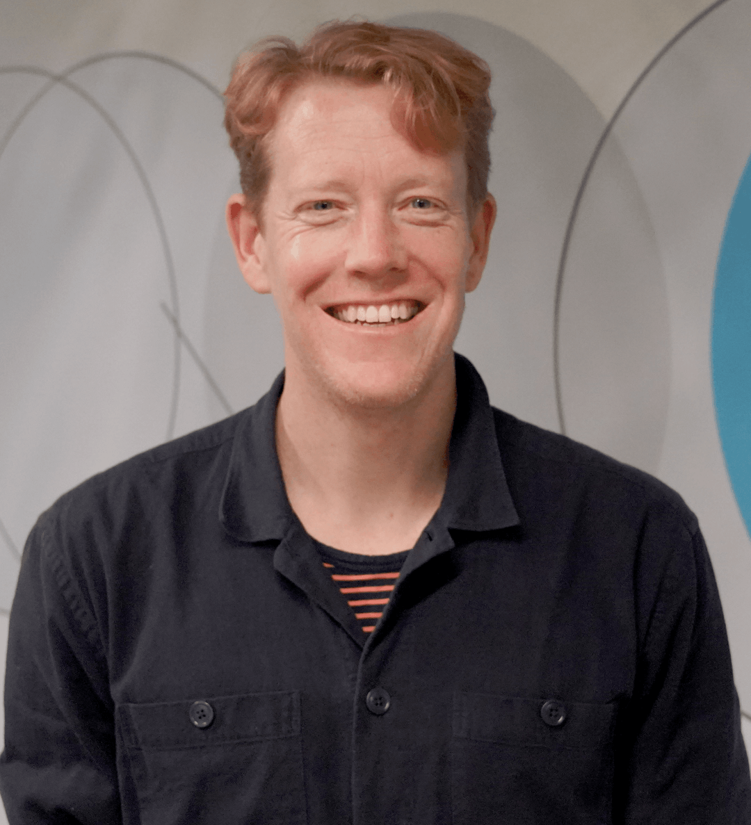 A smiling young man with red hair, wearing a black button-up shirt over a striped t-shirt, standing in front of a gray wall with circular patterns.