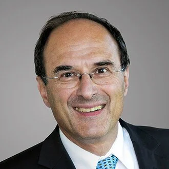 Professional headshot of a middle-aged man with dark hair, wearing glasses, a suit, and a blue polka dot tie, smiling against a gray background.