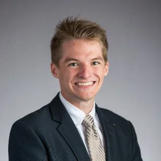 A smiling young man in a dark suit and tie, with a neutral gray background.