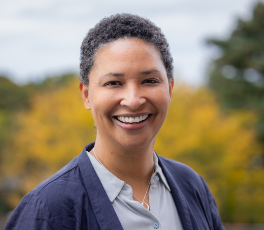 Smiling woman with short curly gray hair outdoors with yellow and green trees in the background.