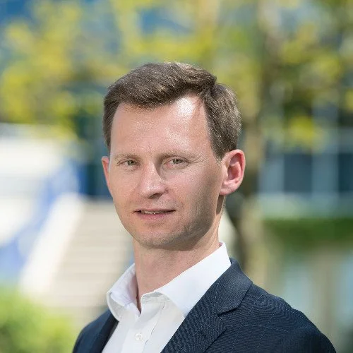 A young man with short brown hair and light skin, wearing a white dress shirt and navy blazer, standing outdoors with blurred trees and building in the background.