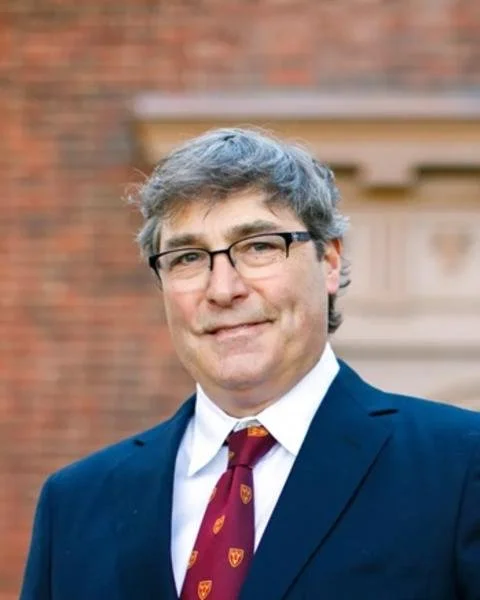 A man with gray hair and glasses wearing a navy suit, white shirt, and maroon tie with a crest, standing outside in front of a brick building.