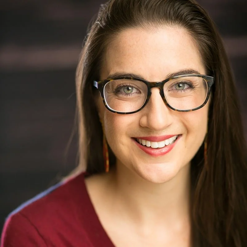 Close-up of a smiling woman with long brown hair, glasses, and earrings, wearing a maroon top against a dark background.