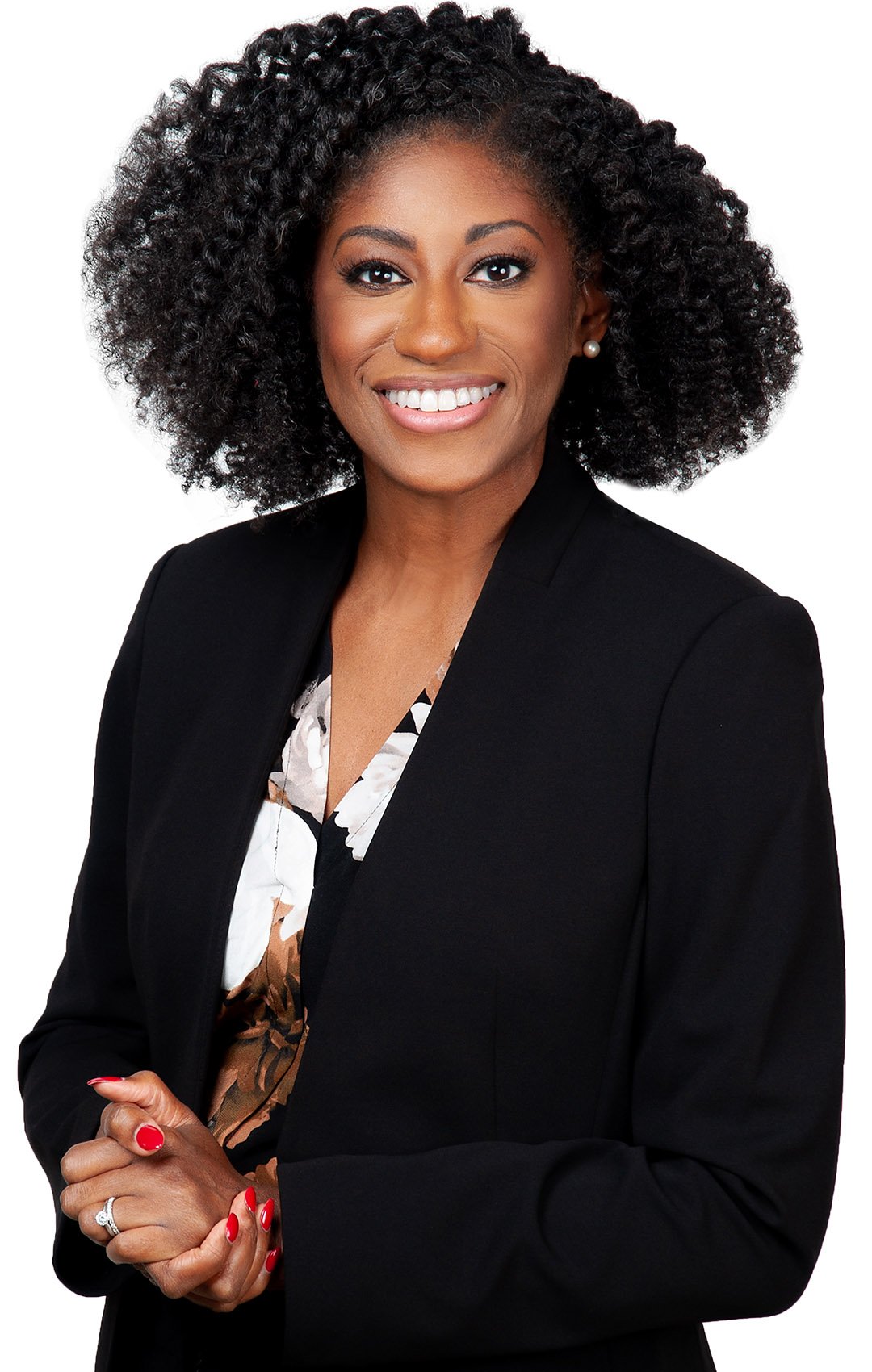 A woman with dark, curly hair wearing a black blazer and a floral blouse, smiling with her hands clasped in front of her.